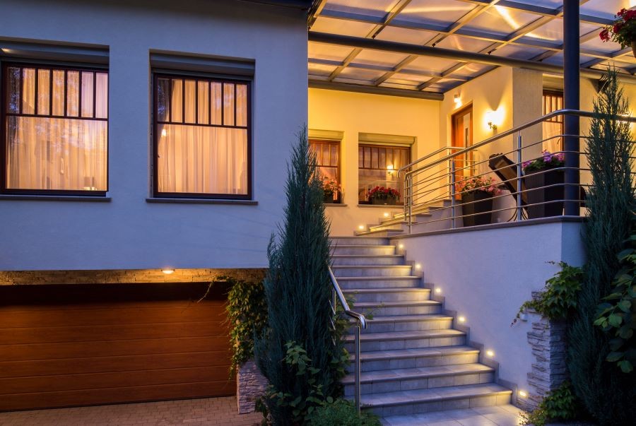 Well-lit stairs surrounded by shrubs lead to a well-lit deck and the front door of a home. 