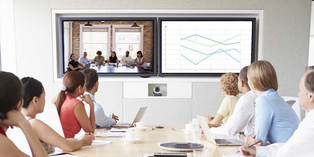 Employees congregate on a wooden boardroom table in front of two displays, one featuring a video call and the other a digital presentation. 