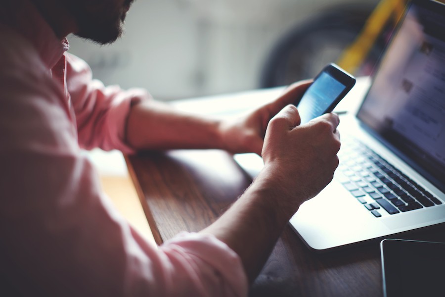 A man sitting on a desk using his phone and laptop.