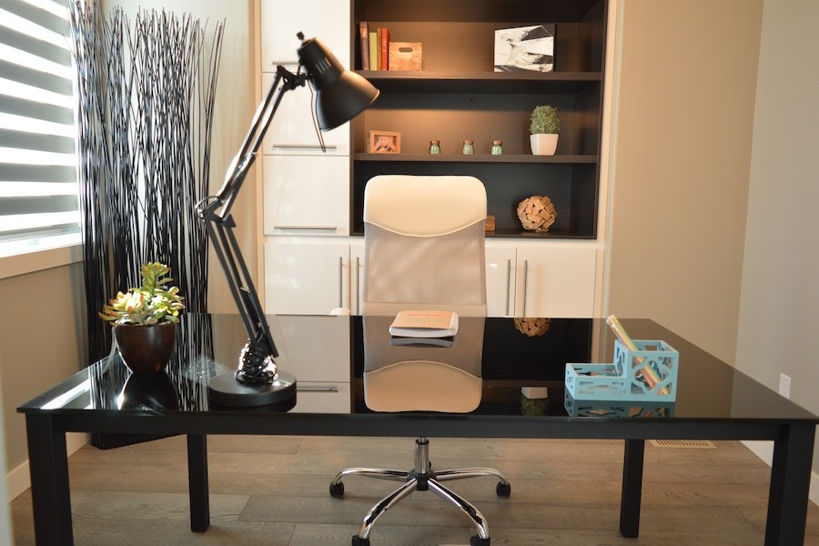 Shot of small home office with a black desk, white chair and white book shelf in the back with a small window and tabletop lamp.