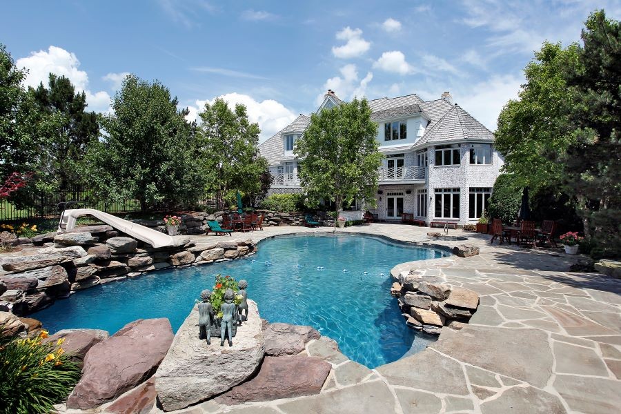  The backyard of a large white home with a pool, slide, and numerous sitting areas.