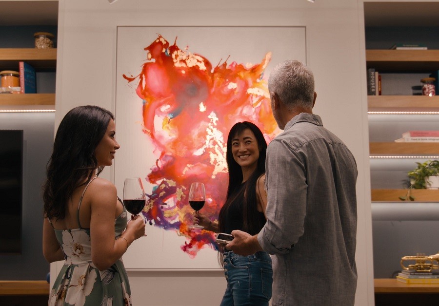 A group of people at a dinner party stand in front of a well-lit piece of artwork. 