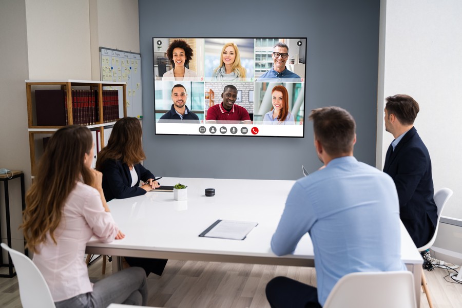 Small conference room with employees at a small desk video conferencing with coworkers shown on a wall-mounted display.