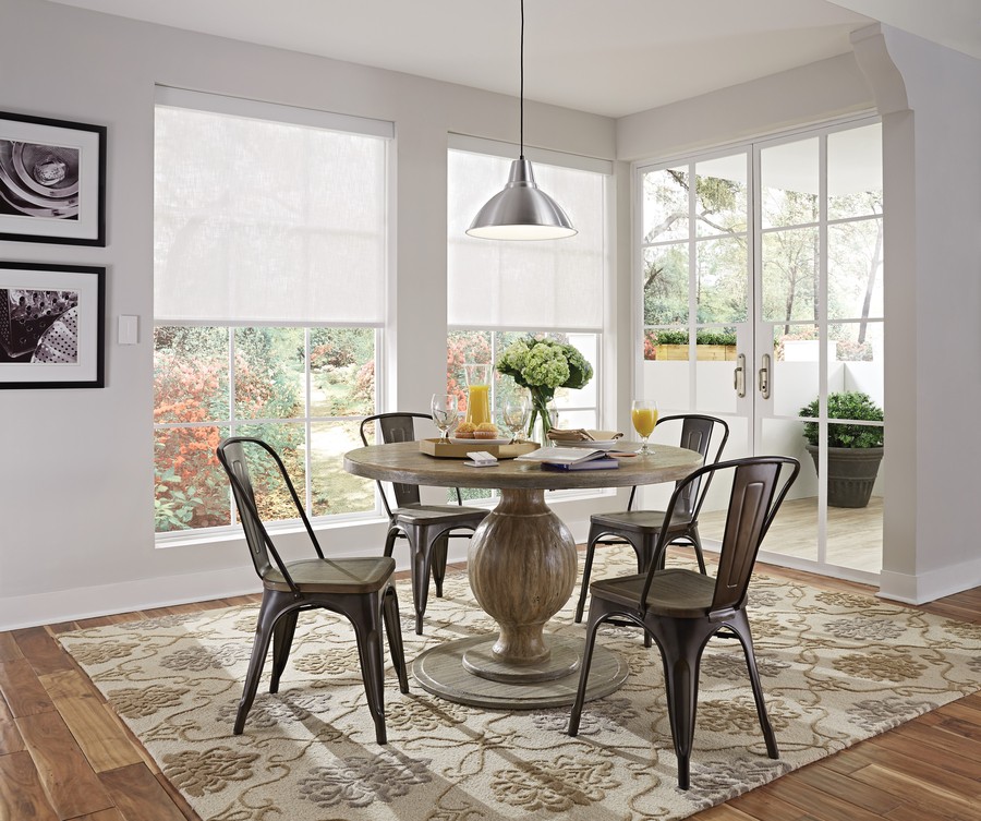 Bright dining area with large windows covered by motorized shades, a round wooden table, metal chairs, and a pendant light.