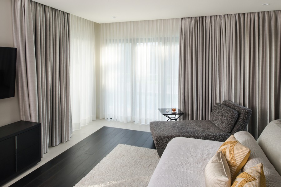 A stylish bedroom featuring a mix of sheer and opaque motorized drapes, contemporary furniture, and a wall-mounted TV.