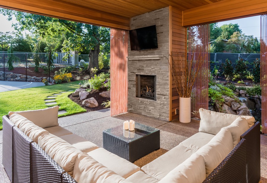 Covered outdoor patio area with fireplace, TV, wicker furniture, and view of landscaped garden.
