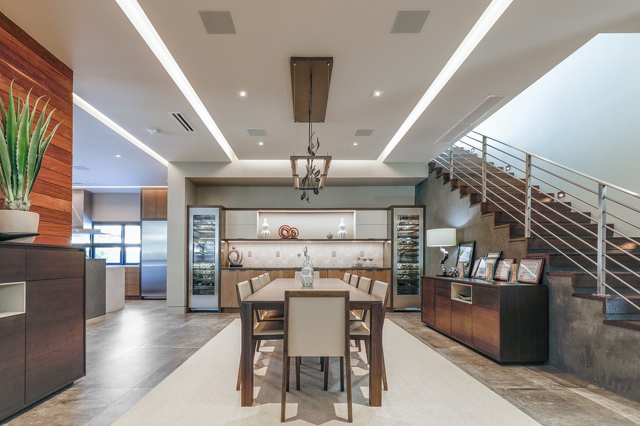 Modern dining room with wooden table, LED lighting, staircase with metal railing, in-ceiling speakers, and an integrated wine refrigerator.
