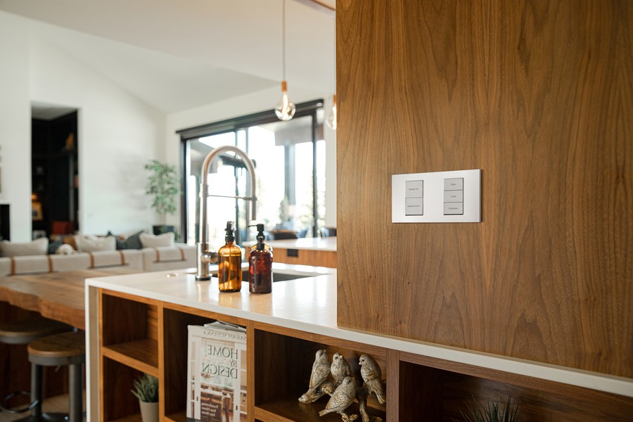 Modern kitchen with smart lighting control panel integrated into a wooden accent wall with sleek buttons for adjusting lighting and shades.